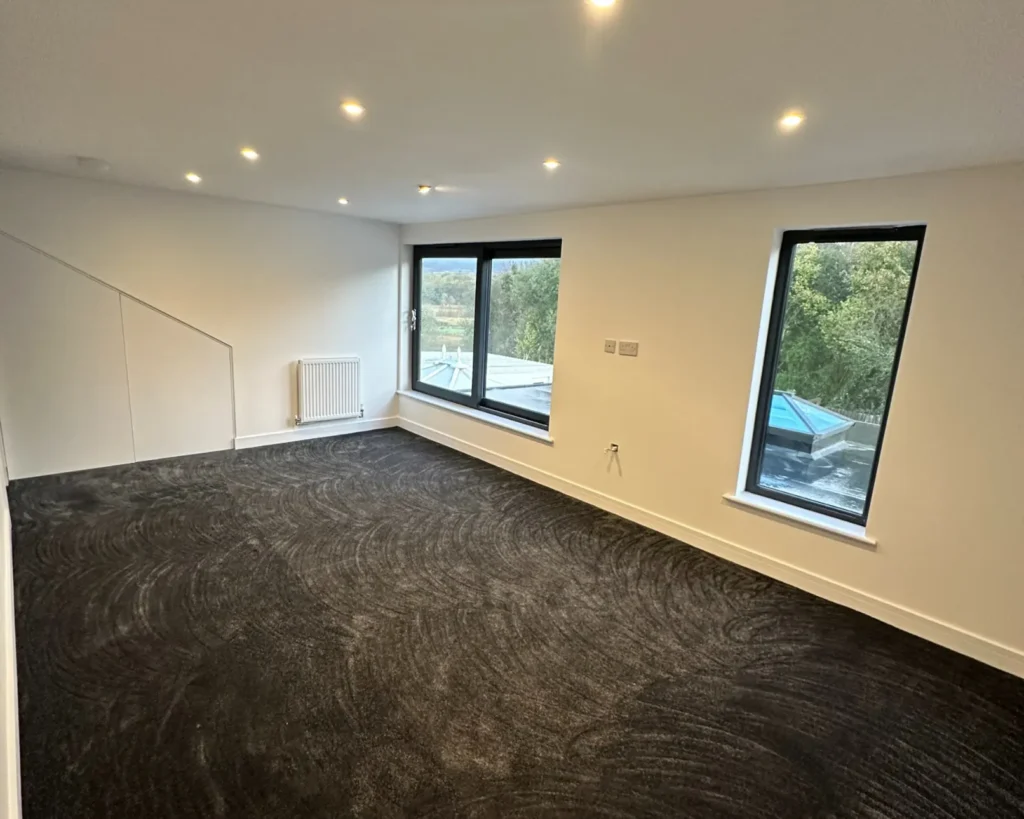 photo of a Loft Conversions in Bridgend & South Wales, spacious loft conversion room with dark carpet, recessed ceiling lights, and large windows overlooking a garden.