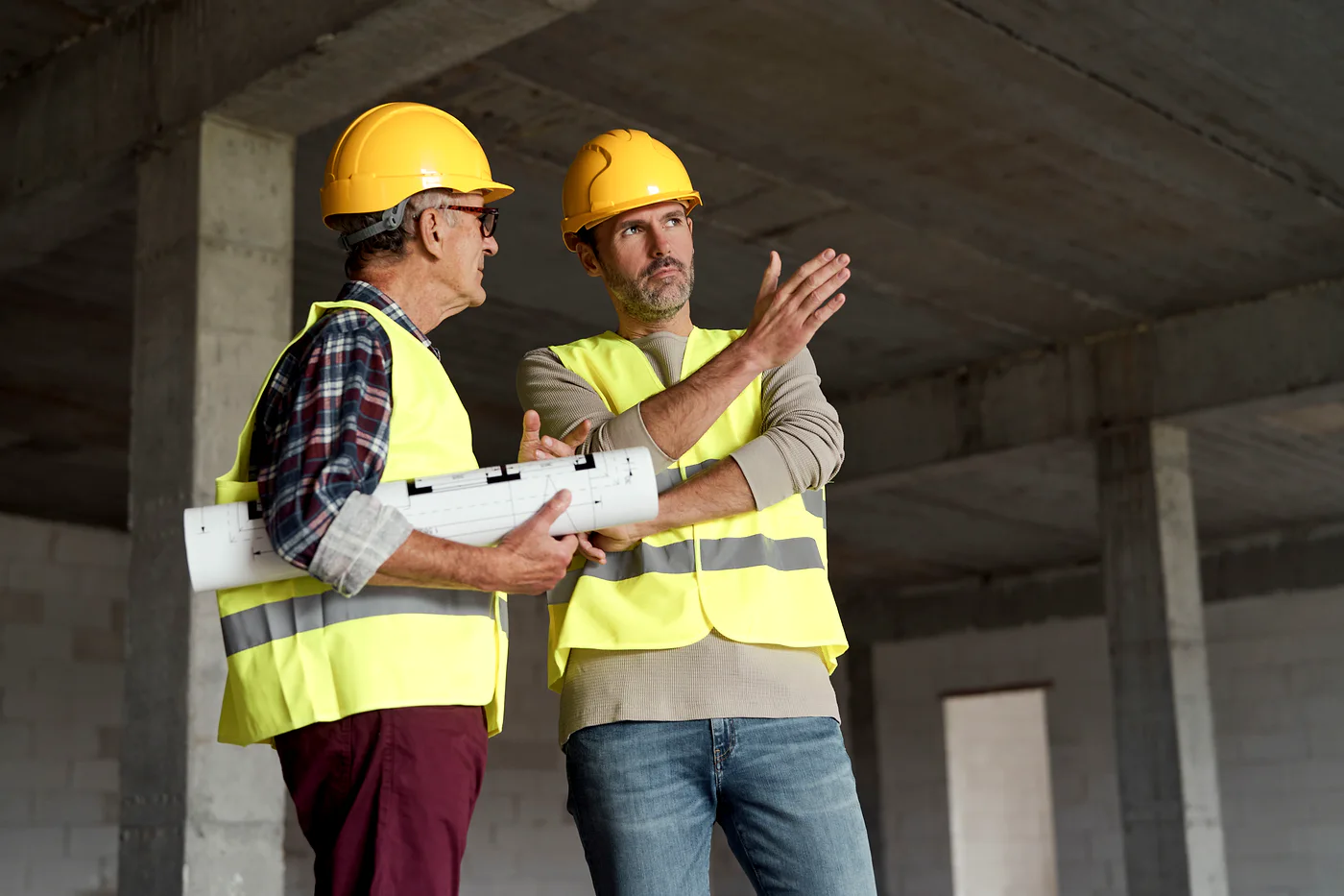 Two construction workers in safety gear discussing a building plan inside an unfinished concrete structure